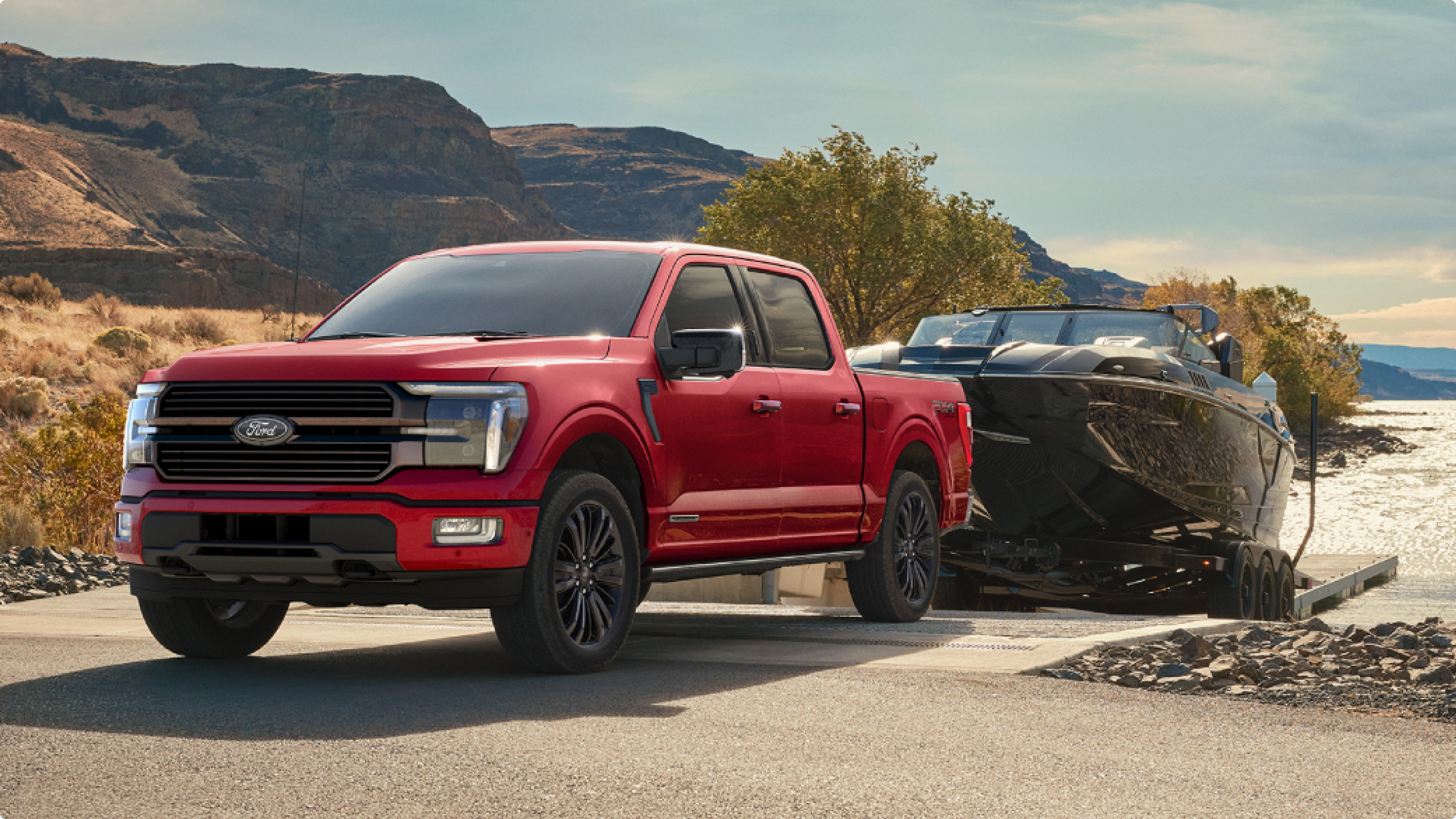 Ford F-150® roja remolcando una lancha negra en una rampa frente al agua con montañas de fondo