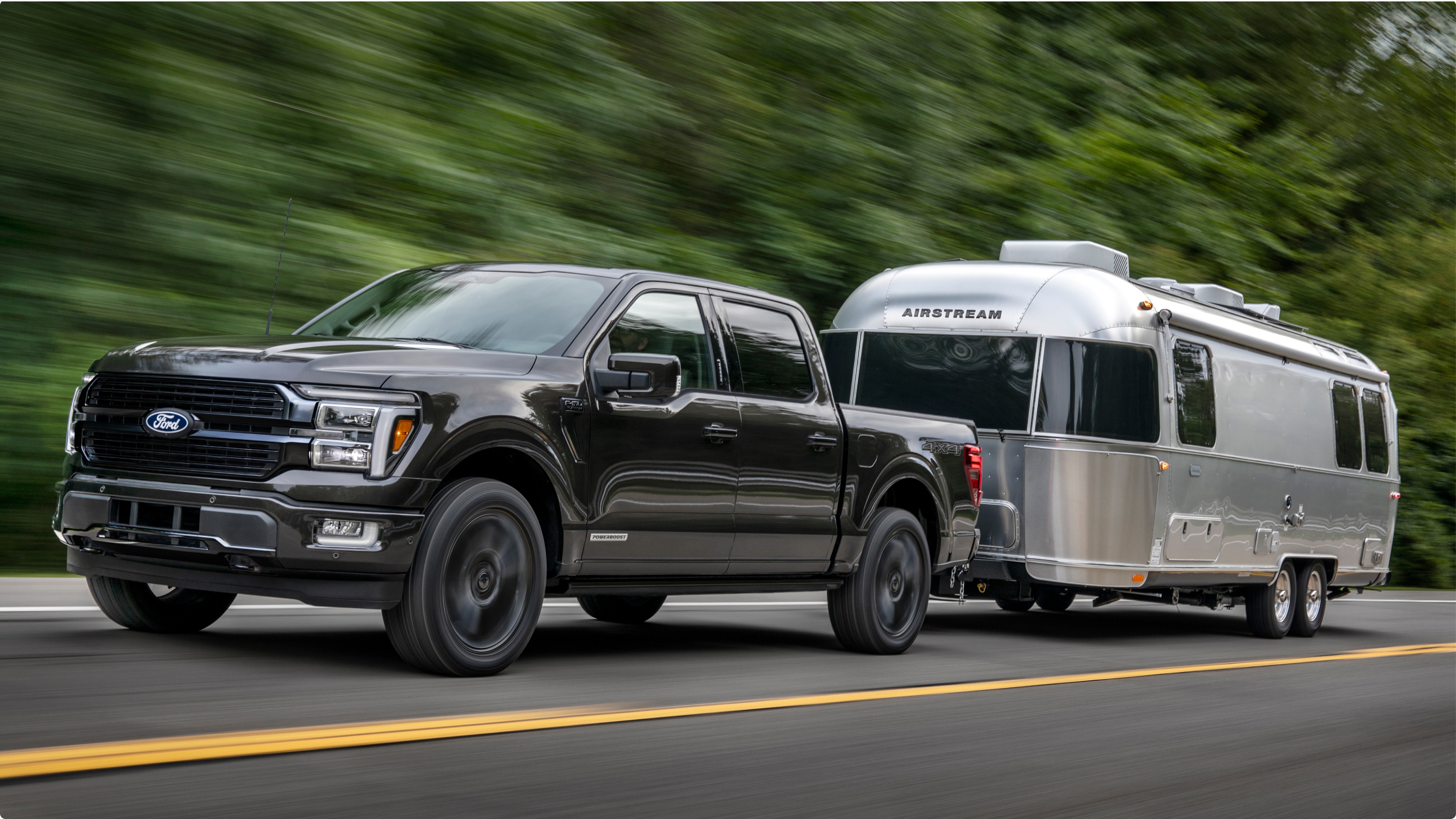 Ford F-150® oscura avanzando por la carretera remolcando un remolque plateado con árboles de fondo