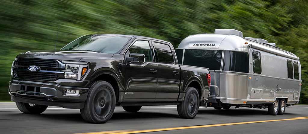 Ford F-150® oscura avanzando por la carretera remolcando un remolque plateado con árboles de fondo